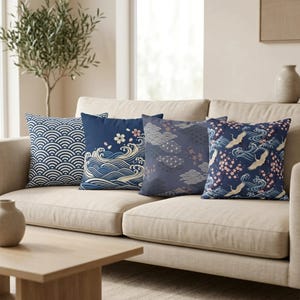 Puede incluir: Cuatro cojines decorativos con diseños de inspiración japonesa descansan sobre un sofá beige. Los cojines presentan patrones de olas, nubes, flores de cerezo y grullas en tonos azules, blancos y rosas. Los cojines son cuadrados.