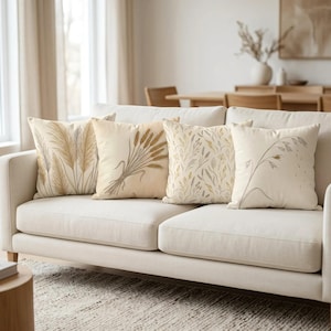 Könnte beinhalten: Vier quadratische Dekokissen auf einem hellen Sofa. Jedes Kissen zeigt eine einzigartige botanische Illustration in Beige-, Sand- und sanften Grautönen, die verschiedene getrocknete Gräser, Weizenähren und zarte Wiesenpflanzenmotive darstellen.