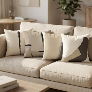 May include: A set of four square throw pillows featuring abstract designs in neutral tones of cream, beige, and dark brown. Each pillow displays unique patterns including vertical lines, horizontal brushstrokes, and layered geometric shapes.