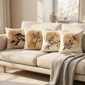 May include: Four decorative throw pillows with Japanese-inspired designs rest on a beige sofa. Each pillow features a different black ink illustration against a tan background: a bonsai tree, bamboo stalks, and flowering branches. A neutral-toned blanket drapes over the sofa.