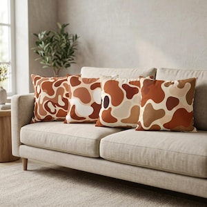 May include: Four square throw pillows with a brown and beige abstract pattern rest on a beige sofa. The pillows have a repeating design of organic shapes in various shades of brown, beige, and white. The sofa sits on a light-colored rug.