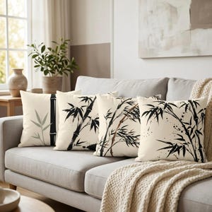 May include: Four decorative throw pillows with bamboo designs. The pillows feature black and green bamboo illustrations on a cream-colored background. The pillows are arranged on a gray sofa with a beige throw blanket.