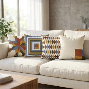 May include: Four square throw pillows arranged on a light-colored sofa. The first features a multi-colored star pattern, the second has concentric squares in blue, yellow, and green, the third displays a chevron pattern, and the fourth is cream with a color-blocked corner.