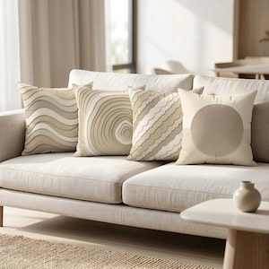May include: Four square throw pillows arranged on a sofa. The pillows feature neutral beige and cream tones with various patterns, including horizontal wavy lines, concentric circles, scalloped edges, and a large solid circle design.