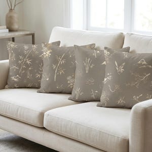 Può includere: Quattro cuscini decorativi quadrati con sfondo color talpa e disegni dorati di ispirazione asiatica. I motivi includono bambù, uccelli e paesaggi montani. I cuscini sono su un divano color crema.
