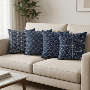 Peut inclure: Quatre coussins carrés disposés sur un canapé beige. Chaque coussin est bleu marine foncé avec un motif géométrique cousu blanc distinct, incluant des grilles triangulaires, des cercles entrelacés, des losanges verticaux et une grande étoile centrale.