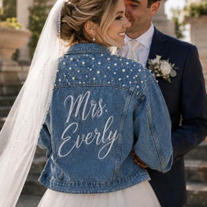 Puede incluir: Una chaqueta de mezclilla azul con adornos de perlas blancas en la parte superior de la espalda y los hombros. La parte posterior de la chaqueta muestra el texto en letra blanca "Mrs. Everly" impreso en una fuente grande y elegante.
