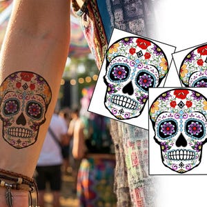 Może przedstawiać: Kolorowy tymczasowy tatuaż z czaszką cukrową jest wyświetlany na ramieniu, z podobnym wzorem na trzech kartach. Czaszka cukrowa ma skomplikowane wzory kwiatowe i geometryczne w kolorach czerwonym, pomarańczowym i fioletowym.
