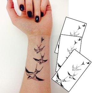 May include: Black and white temporary tattoo of swallows in flight, applied to the wrist. The design features several birds with outstretched wings, creating a sense of movement. Additional tattoo sheets with the same design are visible.