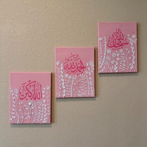 Puede incluir: Tres lienzos rosas con caligrafía árabe en cursiva. Cada lienzo presenta diseños florales blancos en el borde inferior. Los lienzos son rectangulares y están montados en una pared beige.