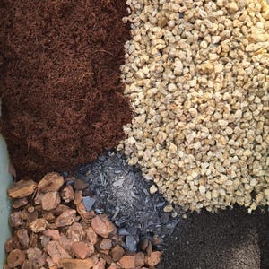 May include: A close-up view of a container divided into sections, each filled with different types of gardening materials. The materials include dark brown mulch, light-colored gravel, dark gray slate, and dark brown soil. The materials are for gardening.