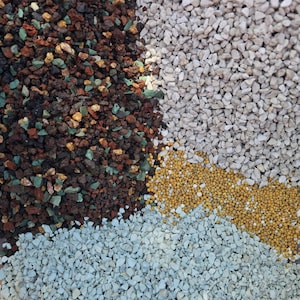 Puede incluir: Una colección de pequeñas piedras decorativas y gránulos dispuestos en cuatro secciones distintas. La parte superior izquierda presenta roca volcánica de color marrón oscuro, negro y verde azulado. La parte superior derecha contiene grava gris claro. La parte inferior derecha tiene gránulos amarillos y la parte inferior izquierda consiste en piedra triturada de color gris azulado claro.