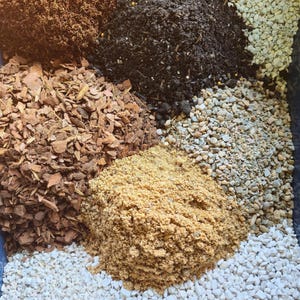 Pode incluir: Uma coleção de seis materiais de jardinagem distintos dispostos em pilhas separadas, incluindo turfa castanho-escura, terra preta para vasos, aparas de madeira bege claro, areia bege grossa, cascalho cinzento e pequenas pedras de perlite branca.
