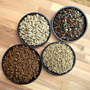 Könnte beinhalten: Vier schwarze, kreisförmige Schalen, gefüllt mit verschiedenen Arten von kleinkörnigem Substratmaterial. Eine Schale enthält hellbeige Kiesel, eine andere dunkelbraune Erdkügelchen, eine dritte feines beiges Granulat und die letzte eine Mischung aus roten, schwarzen und türkisfarbenen Steinen.