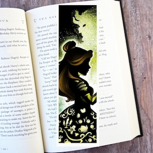 Op de afbeelding: Een zwart-gele boekenlegger met een silhouet van een vrouw in een jurk met decoratieve elementen. De achtergrond toont een silhouet van een beest. De boekenlegger is in een open boek geplaatst.