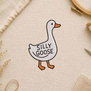 May include: Embroidered patch featuring a white goose with the words "SILLY GOOSE" in black. The goose has an orange beak and feet, with a black outline. The patch is on a beige fabric background.