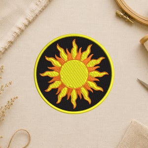 May include: Embroidered patch featuring a stylized sun design. The sun has a yellow center with orange and yellow rays, set against a black background. The patch is circular with a yellow border. The patch is likely for clothing or accessories.