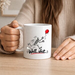 May include: A white ceramic mug featuring a black and white illustration of a skeleton running after a red balloon. Below the skeleton, the text "Well, shit..." is printed in black letters with a red underline, centered on the mug's exterior.