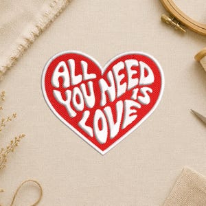 May include: Embroidered patch in the shape of a red heart with the words "ALL YOU NEED IS LOVE" in white, stylized lettering. The patch is on a beige fabric surface, with a wooden embroidery hoop and other craft supplies visible.