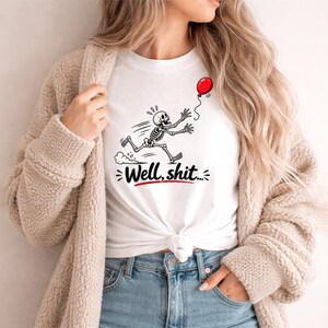 May include: A white t-shirt featuring a black and white graphic of a skeleton running after a red balloon. Below the skeleton, the text "Well, shit..." is printed in a bold black script font with a red underline accenting the phrase.