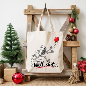 May include: A cream-colored canvas tote bag featuring a black and white graphic of a skeleton running after a red balloon. Below the skeleton, the text "Well, shit..." is printed in black with a red underline accenting the phrase.