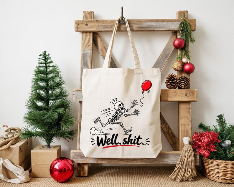 May include: A cream-colored canvas tote bag featuring a black and white graphic of a skeleton running after a red balloon. Below the skeleton, the text "Well, shit..." is printed in black with a red underline accenting the phrase.