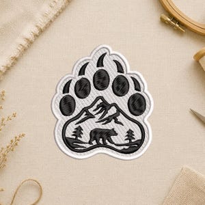 May include: Embroidered patch in the shape of a bear paw, featuring a black bear, mountains, and trees. The design is outlined in white, with black details. This patch is suitable for jackets, bags, or other fabric items.