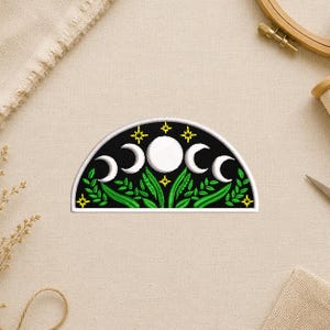 May include: Embroidered patch featuring a crescent moon design with phases of the moon, stars, and green foliage on a black background. The patch has a white border and is in a half-circle shape. Perfect for jackets, bags, or other fabric items.