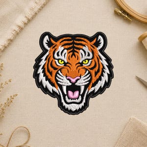 May include: Embroidered tiger head patch with orange, black, white, pink, and yellow details. The tiger is snarling, with its mouth open, teeth bared, and eyes narrowed. The patch has a black border and is set against a neutral background.