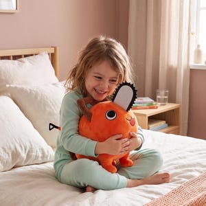K&ouml;nnte beinhalten: Ein orangefarbenes Pl&uuml;schtier in Form einer Kettens&auml;ge mit einer schwarz-wei&szlig;en Klinge und braunen Griffen. Das Spielzeug wird von einer Person in hellgr&uuml;ner Pyjama gehalten. Der Hintergrund umfasst ein Bett, Kissen und einen Nachttisch.