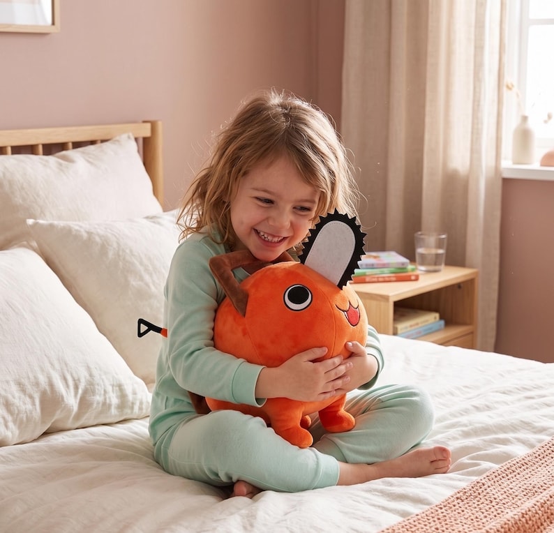 K&ouml;nnte beinhalten: Ein orangefarbenes Pl&uuml;schtier in Form einer Kettens&auml;ge mit einer schwarz-wei&szlig;en Klinge und braunen Griffen. Das Spielzeug wird von einer Person in hellgr&uuml;ner Pyjama gehalten. Der Hintergrund umfasst ein Bett, Kissen und einen Nachttisch.