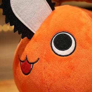 K&ouml;nnte beinhalten: Nahaufnahme eines orangefarbenen Pl&uuml;schtiers mit einem s&auml;gekettenartigen Kopfst&uuml;ck. Das Spielzeug hat ein gro&szlig;es schwarz-wei&szlig;es Auge, einen roten Mund und einen l&auml;chelnden Ausdruck. Das Pl&uuml;schtier ist weich und flauschig.