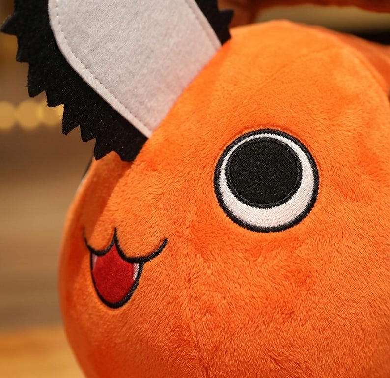 K&ouml;nnte beinhalten: Nahaufnahme eines orangefarbenen Pl&uuml;schtiers mit einem s&auml;gekettenartigen Kopfst&uuml;ck. Das Spielzeug hat ein gro&szlig;es schwarz-wei&szlig;es Auge, einen roten Mund und einen l&auml;chelnden Ausdruck. Das Pl&uuml;schtier ist weich und flauschig.