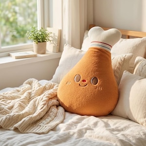 May include: A novelty chicken leg-shaped cushion in orange, featuring an embroidered smiling face and a white chef's hat. The cushion is placed on a bed with white pillows and a cream cable-knit throw. The scene is lit by natural light.