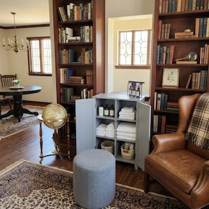 May include: A light gray storage cabinet with two open doors, featuring four shelves filled with white folded towels, small bottles, and two woven baskets. The cabinet is positioned between two tall wooden bookshelves in a room with a leather chair.