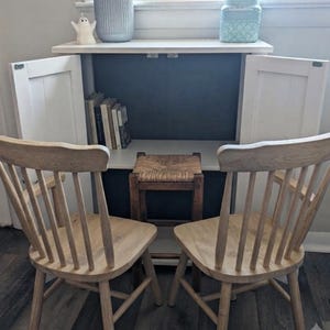 May include: A white cabinet with two open doors featuring a dark interior, a shelf holding several books, and a small wooden stool with a woven seat. Two light-colored wooden spindle chairs are positioned in front of the cabinet.