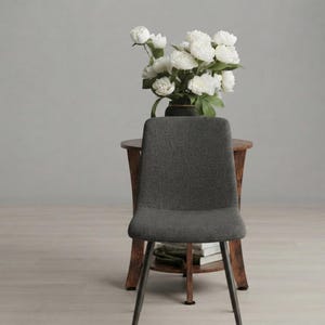 May include: A dark gray upholstered chair with a high back and four thin black legs. Behind the chair sits a small wooden side table with a curved base, holding a dark vase filled with a large bouquet of white flowers and green leaves.