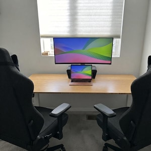 May include: A light-colored wooden desk featuring a laptop and a large ultrawide monitor displaying a colorful abstract wallpaper, positioned between two dark gray ergonomic office chairs with armrests in a bright room with a window.