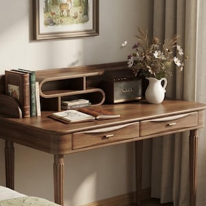 May include: A dark wood desk with two drawers featuring gold-toned handles, a curved open shelf unit, a rectangular speaker, a white ceramic pitcher filled with dried flowers, and an open book. A framed illustration hangs on the wall above.