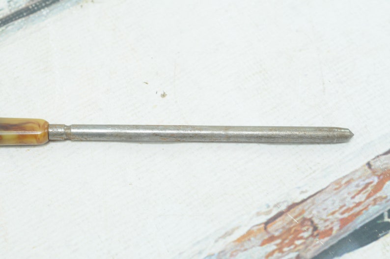 Op de afbeelding: Een metalen gereedschap met een bruin en wit marmerachtig kunststof handvat. Het gereedschap is lang en dun, met een puntig uiteinde.
