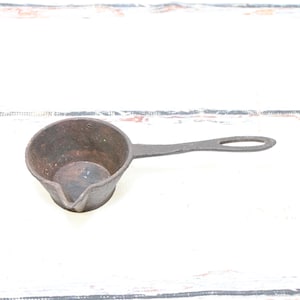 May include: A small, antique cast iron ladle with a long handle and a pouring spout. The ladle is dark brown with signs of wear and age, suggesting it's a vintage kitchen tool. The handle has a hole for hanging.