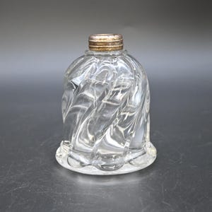 May include: A clear glass bottle with a distinct spiral swirl pattern throughout its body. The bottle features a rounded, bell-like shape and is topped with a small, metallic, threaded cap that has a weathered, aged finish on its surface.