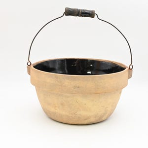 May include: A tan-colored, ceramic bucket with a black interior. It has a metal handle with a wooden grip. The bucket is round with a slightly flared rim.