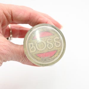 以下が含まれることがあります： 金色の「BOSS」の文字がピンクと金の背景に配置された透明な丸いオブジェクト。白い背景の中で、手で持たれています。