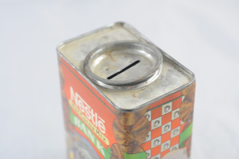 Pode incluir: Um cofre de metal vintage em forma de caixa de leite de chocolate Nestl&eacute;. A caixa &eacute; vermelha e branca com uma imagem de uma caixa de leite de chocolate e as palavras "Nestle's Chocolate Milk" na frente. O cofre tem uma fenda no topo para moedas.