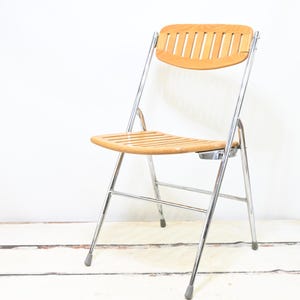 May include: A folding chair with a light brown wooden seat and backrest. The backrest has vertical slats. The frame is made of shiny, silver-colored metal. The chair is set against a white background.