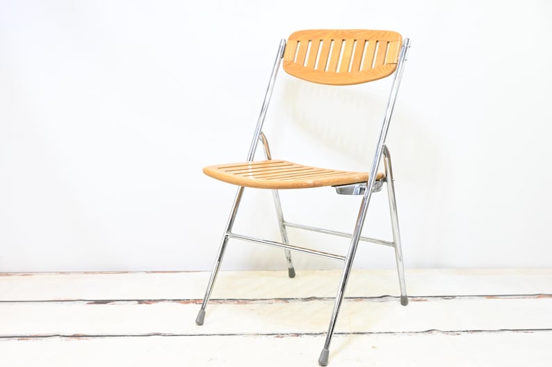 May include: A folding chair with a light brown wooden seat and backrest. The backrest has vertical slats. The frame is made of shiny, silver-colored metal. The chair is set against a white background.