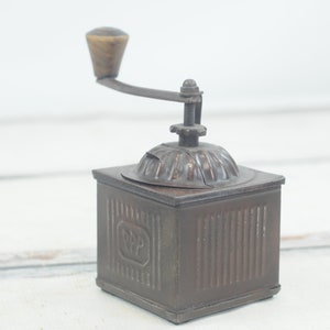 Könnte beinhalten: Eine antike, handbetriebene Kaffeemühle aus Metall mit einem Holzgriff. Die Mühle hat einen quadratischen Sockel mit einem gerippten Design und die Buchstaben "R.P.P." sind auf der Vorderseite eingeprägt.