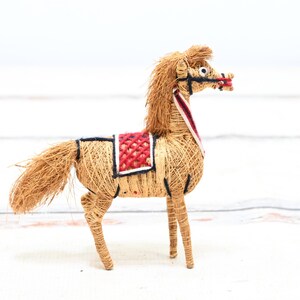 May include: A handmade horse figurine made of straw with a red and white saddle. The horse has a long mane and tail and is standing on a white surface.