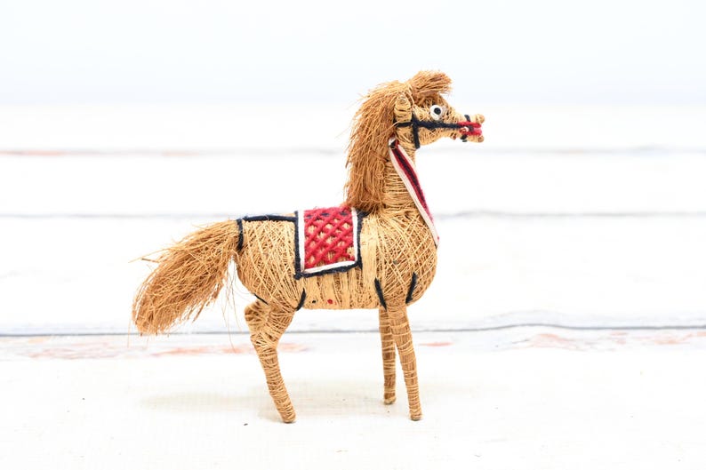May include: A handmade horse figurine made of straw with a red and white saddle. The horse has a long mane and tail and is standing on a white surface.